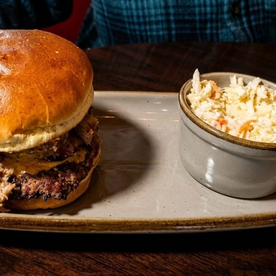 Pulled pork and beef burger with melted cheese served alongside creamy coleslaw on a ceramic platter.