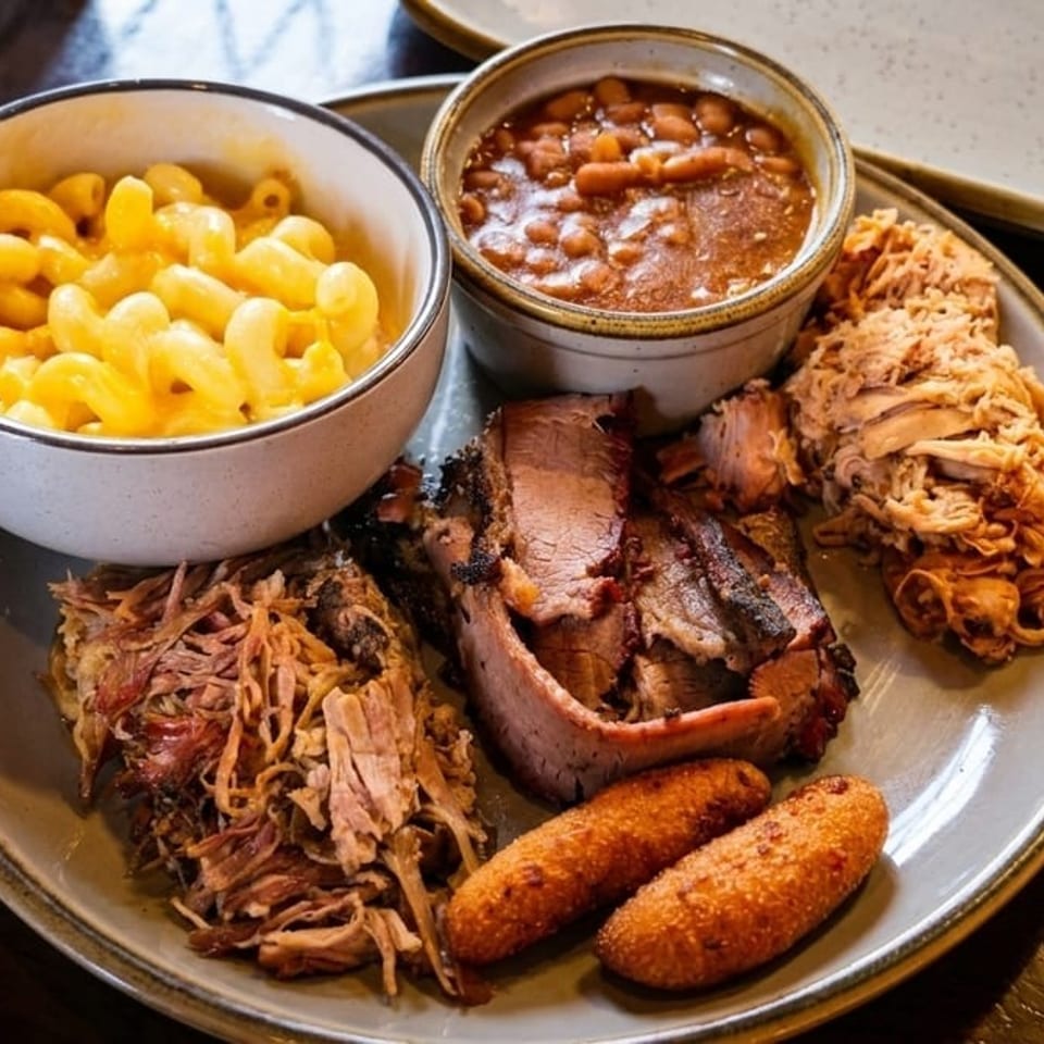 Barbecue platter with pulled pork, sliced brisket, baked beans, macaroni and cheese, and fried sides on a wooden table.