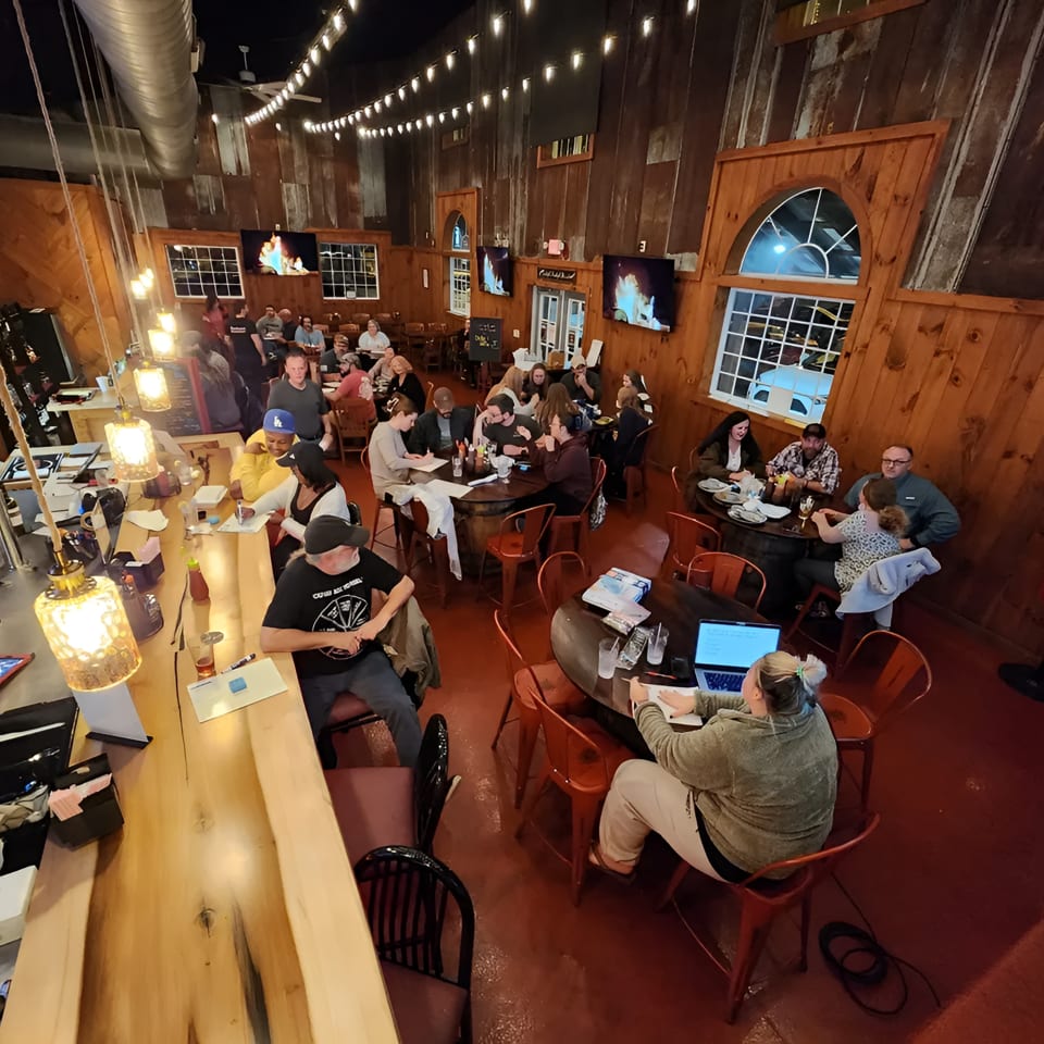 Diners and staff fill a rustic restaurant with wooden walls, string lights, and a long bar.