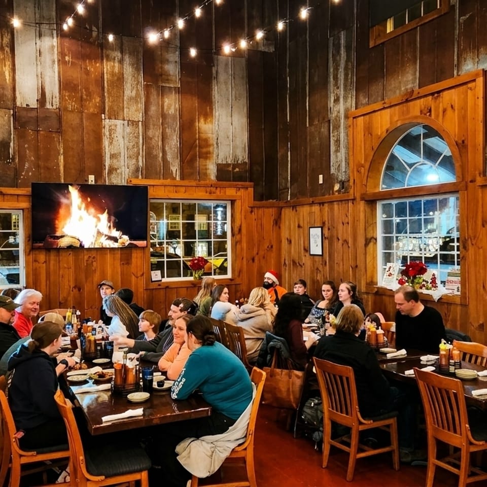 Diners seated at wooden tables in a rustic restaurant with string lights and fireplace videos on TVs.