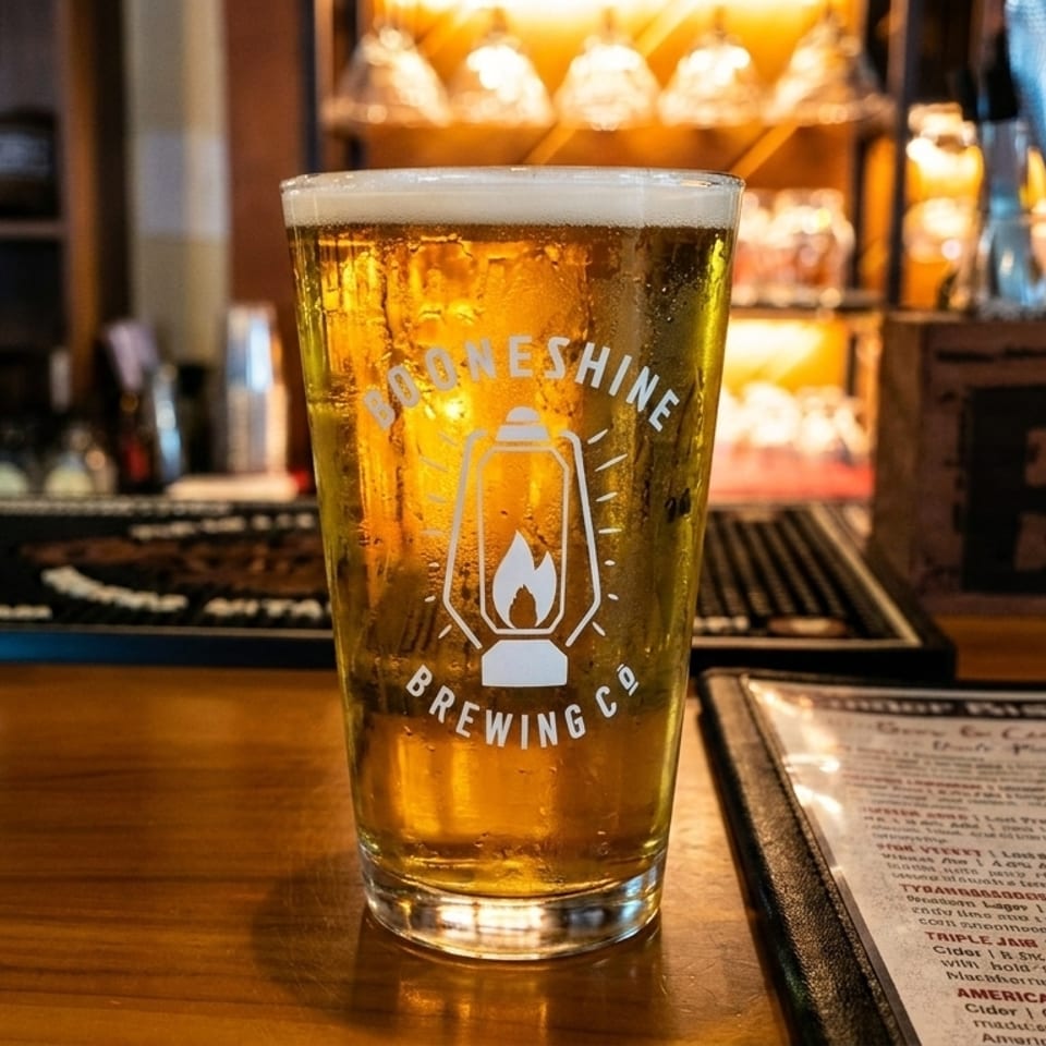 A pint glass of Booneshine Brewing Co. beer sits on a wooden bar next to a menu.