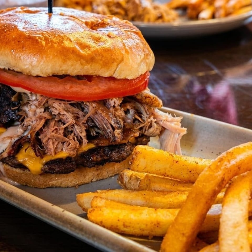 Pulled pork and cheeseburger topped with tomato on a toasted bun, served with seasoned fries on a plate.